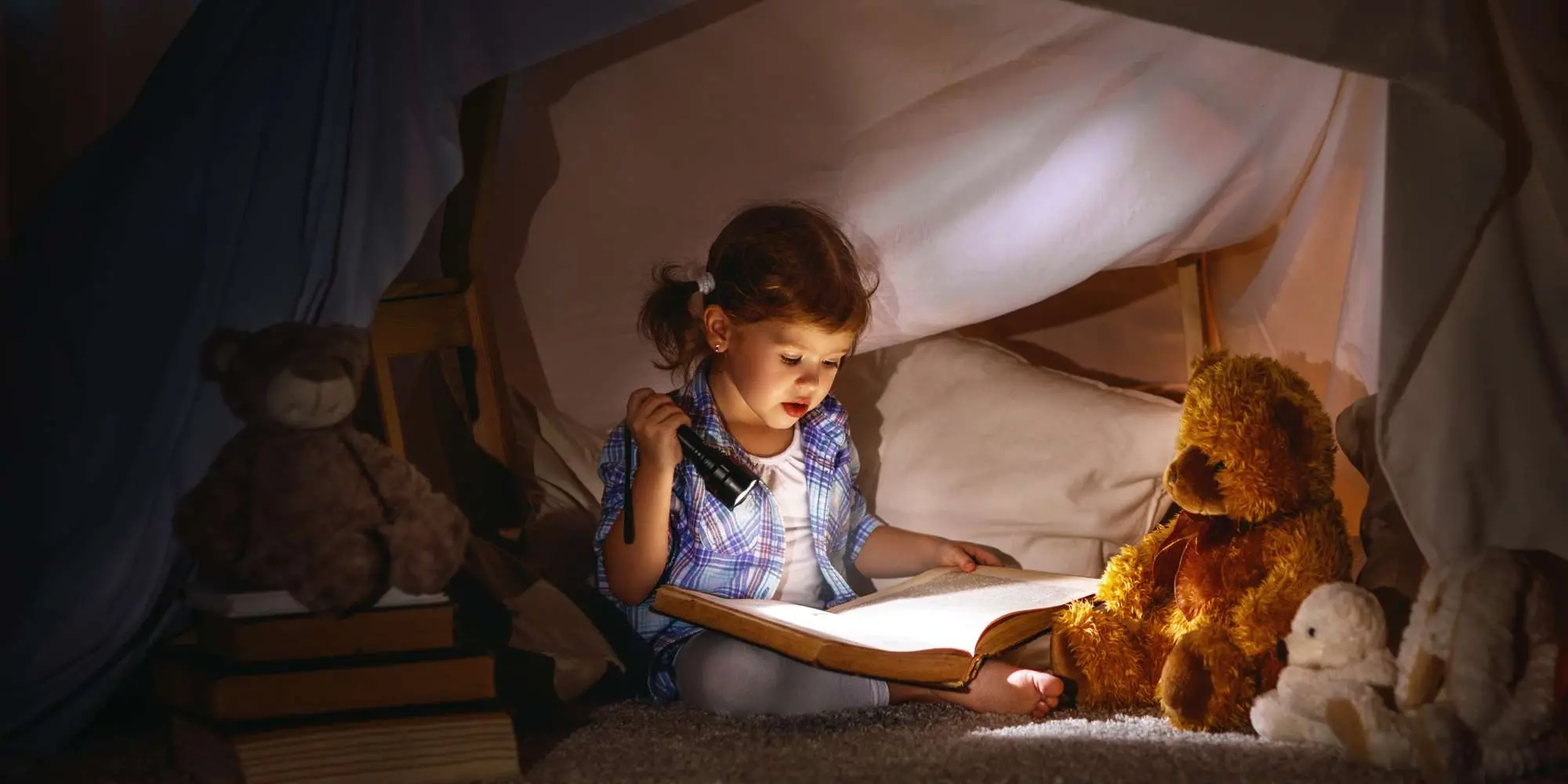 Liten tjej läser bok med ficklampa i en koja inomhus.